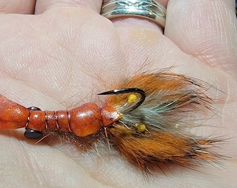 May include: A close-up of a hand holding a brown and orange fly fishing lure with a black hook and yellow beads. The lure has a fuzzy tail and a segmented body.
