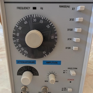 Op de afbeelding: Een lichtgrijze audiogenerator model TAG-101 met een frequentiebereik van 10Hz-1MHz. Het voorpaneel is voorzien van een grote ronde frequentieknop, vijf bereikselectieknoppen, een verzwakkingsknop, een amplitudeknop, een golfvormknop en een aan-uit schakelaar.