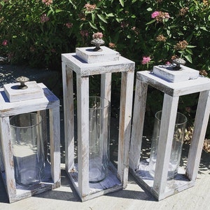 Könnte beinhalten: Drei weiße Holz-Kerzenlaternen mit Glaszylindern. Die Laternen haben eine abgewetzte weiße Farbe und Metallgriffe.