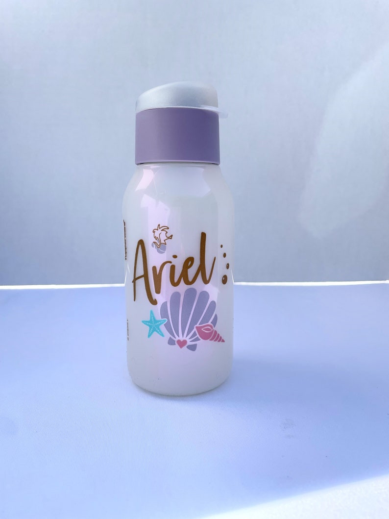 May include: White water bottle with a purple lid and a gold design that says "Ariel" with a seashell and starfish.