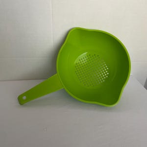 May include: A bright lime green plastic colander featuring a round bowl with a perforated base for draining liquids. The kitchen tool includes a long, flat handle with a circular hole at the end for hanging and two small pouring spouts on the rim.