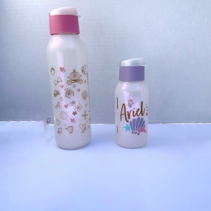 May include: Two white water bottles with pink and purple lids. The larger bottle has a gold design with hearts, flowers, and a carriage. The smaller bottle has a gold design with a seashell, a star, and the word "Ariel".