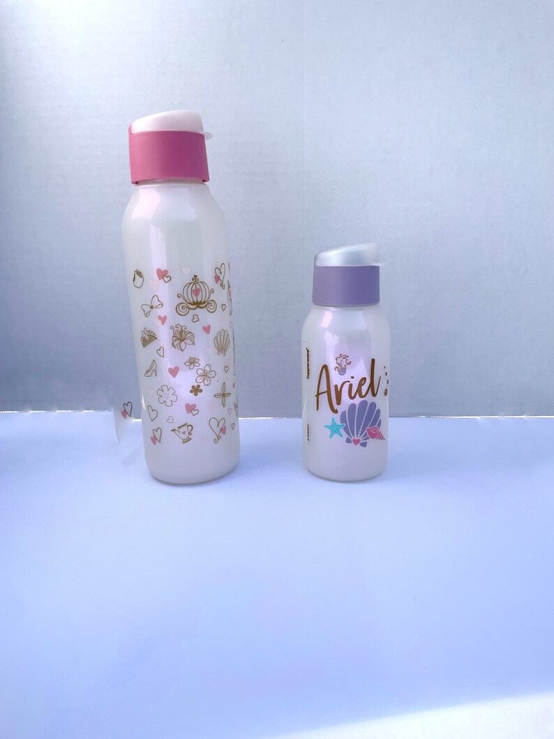 May include: Two white water bottles with pink and purple lids. The larger bottle has a gold design with hearts, flowers, and a carriage. The smaller bottle has a gold design with a seashell, a star, and the word "Ariel".