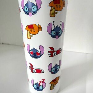 May include: White travel mug with a blue lid and a repeating pattern of Stitch from Lilo & Stitch. Stitch is shown in different expressions, including winking, hearts for eyes, and sleeping. The mug also features a red and orange space ship and a red and orange laser gun.