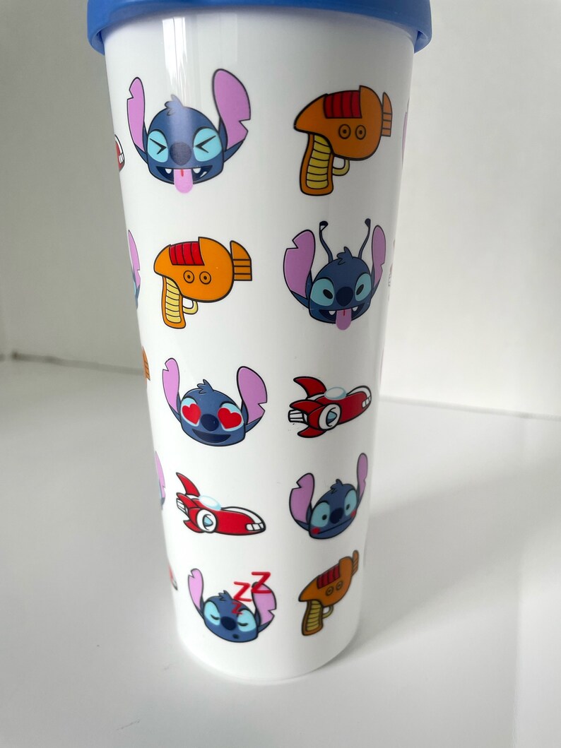 May include: White travel mug with a blue lid and a repeating pattern of Stitch from Lilo & Stitch. Stitch is shown in different expressions, including winking, hearts for eyes, and sleeping. The mug also features a red and orange space ship and a red and orange laser gun.