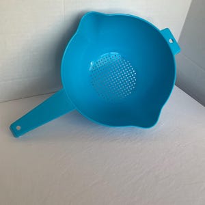 May include: A bright turquoise plastic colander with a handle and a perforated bottom. The colander has a wide bowl shape with a pouring spout and a small hole on the handle for hanging. Kitchen utensil.