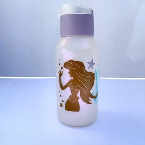 May include: A white water bottle with a purple lid and a gold silhouette of Ariel the Little Mermaid, seaweed, and a starfish on the front.
