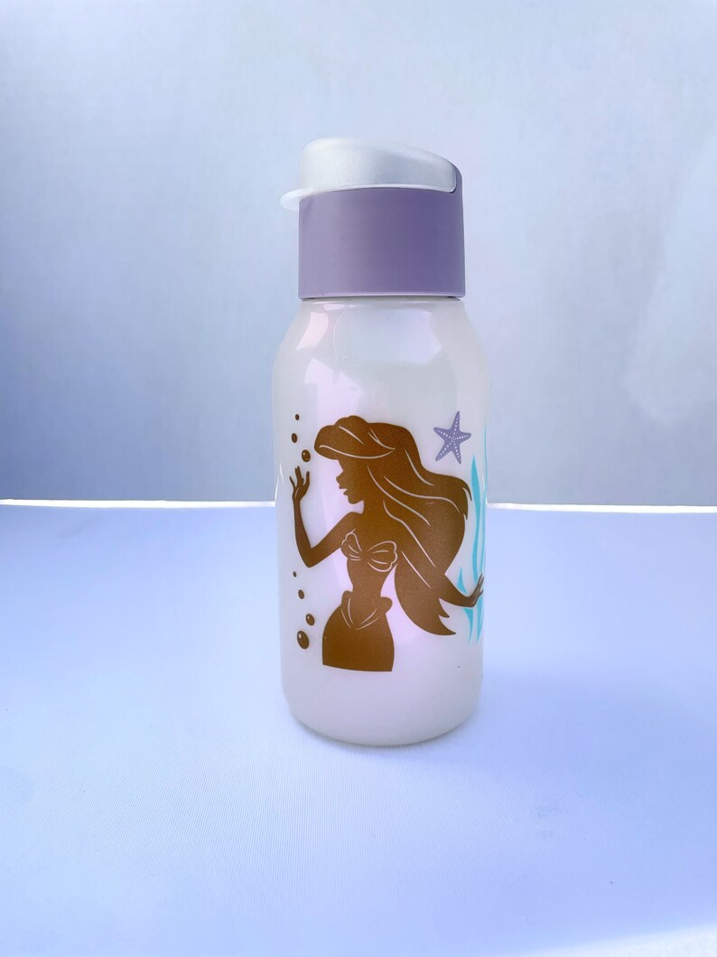 May include: A white water bottle with a purple lid and a gold silhouette of Ariel the Little Mermaid, seaweed, and a starfish on the front.