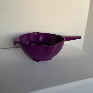 May include: A deep purple plastic mixing bowl with a handle and a pouring spout. The bowl has a scalloped edge and two small handles on either side. The handle is rectangular and extends from the side of the bowl.