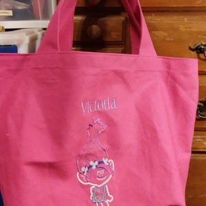 Può includere: Una borsa tote rosa acceso con due manici. La borsa presenta un'immagine ricamata di un personaggio dei cartoni animati e il nome "Victoria" in filo blu chiaro. La borsa è realizzata in un tessuto resistente.