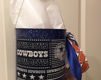 May include: A blue metal bucket with a white handle and a Cowboys theme. The bucket has a white star and the words "Cowboys" repeated three times. The bucket is decorated with white stars and a blue and white ribbon.