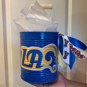 May include: A blue metal can with a white handle and a Los Angeles Rams logo. The can is decorated with blue glitter and has a yellow and blue ribbon attached to it.