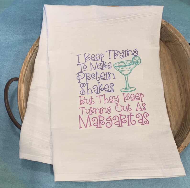 Puede incluir: Toalla de cocina blanca con un dicho humor&iacute;stico sobre hacer batidos de prote&iacute;nas que siempre terminan siendo margaritas. El texto est&aacute; en morado y turquesa, con una ilustraci&oacute;n de un vaso de margarita turquesa.