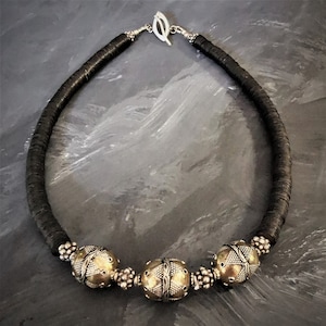 Puede incluir: Un collar de cuero negro con tres cuentas de plata y oro. Las cuentas tienen diseños intrincados.