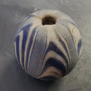 Könnte beinhalten: Eine runde Perle mit einem Loch, verziert mit einem blau-weiß gestreiften Muster. Die Perle hat ein marmoriertes Aussehen mit Wirbeln in Blau, Weiß und Beige. Sie scheint aus Stein oder Glas zu bestehen.