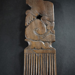 Könnte beinhalten: Ein geschnitzter Holzkamm mit detailliertem Design. Der Kamm zeigt eine schweineähnliche Kreatur auf einem wirbelnden Wolkenmotiv. Die Zähne des Kamms sind gleichmäßig verteilt und verlaufen nach unten. Das Holz hat einen warmen, braunen Farbton.