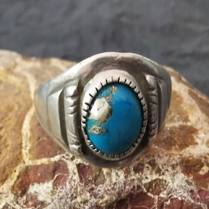 Puede incluir: Un anillo de plata con una gran piedra turquesa ovalada. La piedra tiene un aspecto moteado con tonos azules y dorados. El anillo tiene una banda gruesa y elementos decorativos en los laterales.