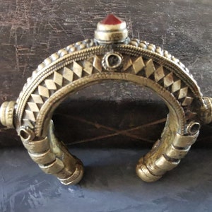 Può includere: Un braccialetto in metallo dorato con un design geometrico e un accento di pietra rossa. Il braccialetto è aperto e ha una forma curva.