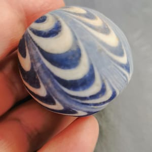 Könnte beinhalten: Ein runder, blau-weißer dekorativer Stein mit einem Wirbelmuster. Der Stein hat eine polierte Oberfläche und wird in einer Hand gehalten. Das Design zeigt abwechselnde Bänder in Weiß und Dunkelblau.