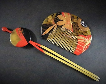 Op de afbeelding: Een decoratieve haar kam en haarspeld set. De kam heeft een waaier vorm met gouden, rode en zwarte bloemmotieven. De haarspeld heeft een ronde, rode, zwarte en gouden kop met een lange, geel en rode stok.
