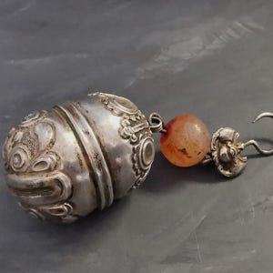 May include: A silver-toned metal pendant featuring an oval-shaped, embossed container with intricate swirling patterns. Attached to the side is a single, round, translucent orange bead followed by a small decorative metal element and a curved hook.
