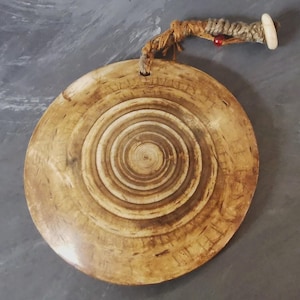 Puede incluir: Un colgante circular de madera de color tostado con un patrón de grano en espiral natural de color marrón oscuro. El colgante está suspendido por un cordón marrón grueso y texturizado, adornado con una pequeña cuenta roja y un cierre cilíndrico blanco.