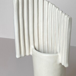 Pu&ograve; includere: Un vaso in ceramica bianca dal design unico. Presenta una base cilindrica e una parte superiore curva a forma di ventaglio con scanalature verticali. La forma moderna e minimalista lo rende adatto all'arredamento.