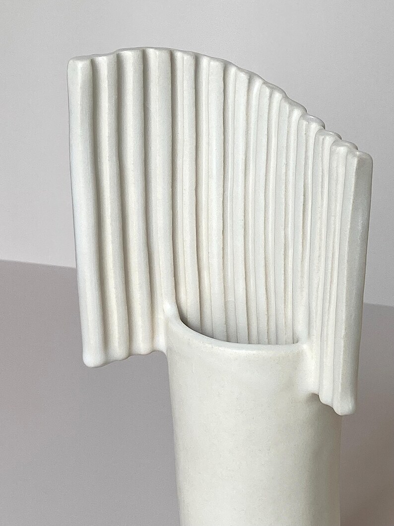 Pu&ograve; includere: Un vaso in ceramica bianca dal design unico. Presenta una base cilindrica e una parte superiore curva a forma di ventaglio con scanalature verticali. La forma moderna e minimalista lo rende adatto all'arredamento.