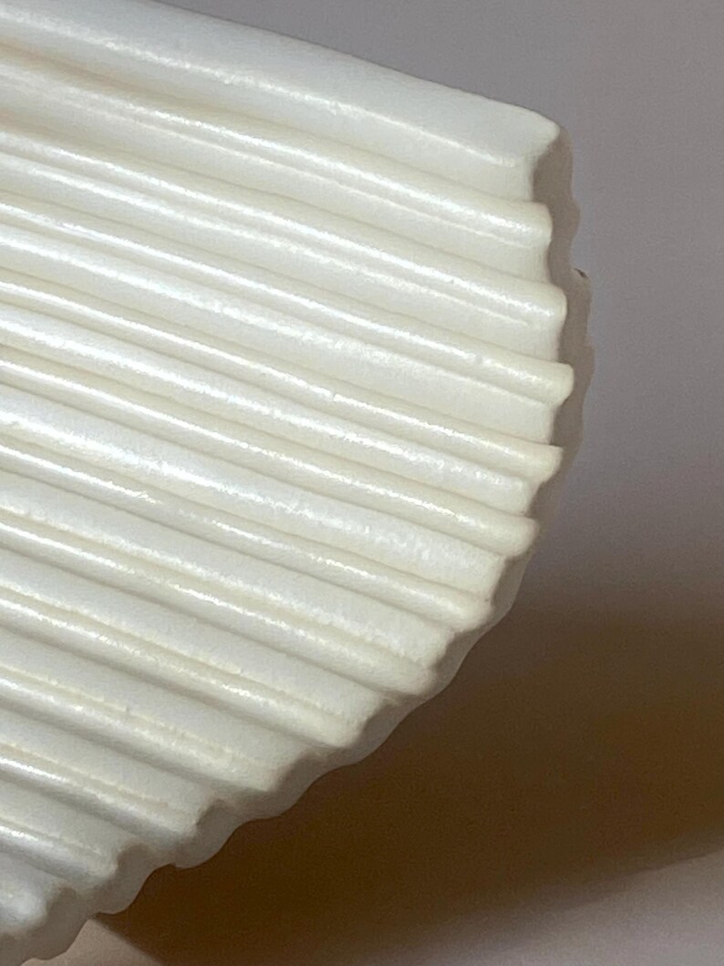 Pu&ograve; includere: Primo piano di un oggetto bianco a forma di ventaglio con una texture a coste. L'oggetto ha un bordo curvo e sembra essere fatto di un materiale solido e opaco. Le creste corrono parallele tra loro, creando un motivo.