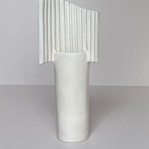 Pu&ograve; includere: Un vaso in ceramica bianca dal design unico. Il vaso ha una base cilindrica e una parte superiore rettangolare con scanalature verticali. La parte superiore &egrave; leggermente angolata, aggiungendo un tocco moderno. Il vaso &egrave; di colore bianco pieno.