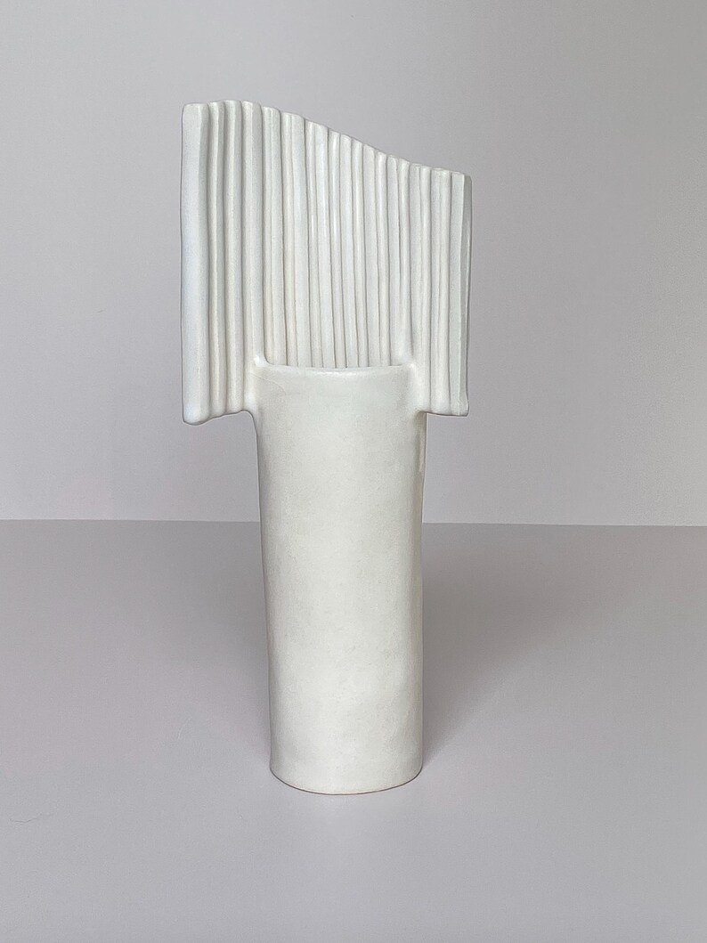 Pu&ograve; includere: Un vaso in ceramica bianca dal design unico. Il vaso ha una base cilindrica e una parte superiore rettangolare con scanalature verticali. La parte superiore &egrave; leggermente angolata, aggiungendo un tocco moderno. Il vaso &egrave; di colore bianco pieno.