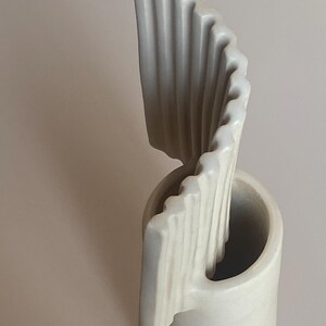 Pu&ograve; includere: Una scultura astratta in ceramica color crema. Il pezzo presenta una base cilindrica con una forma curva a onda che si alza. La superficie strutturata presenta delle scanalature verticali, creando un design dinamico. Scultura decorativa.