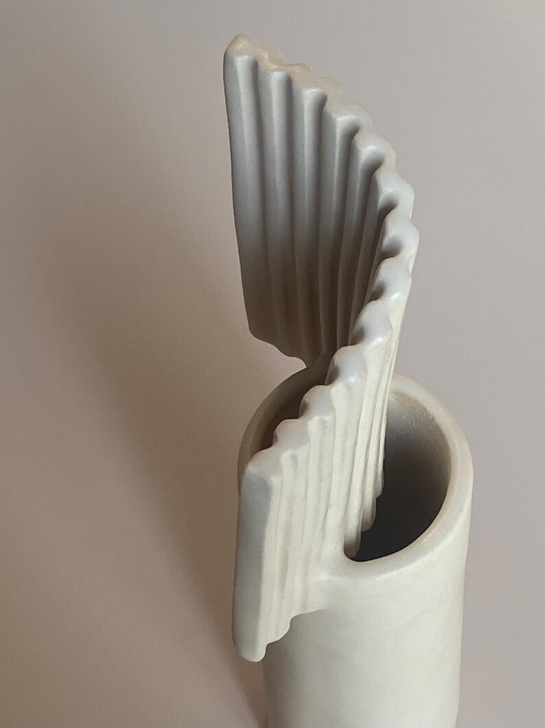 Pu&ograve; includere: Una scultura astratta in ceramica color crema. Il pezzo presenta una base cilindrica con una forma curva a onda che si alza. La superficie strutturata presenta delle scanalature verticali, creando un design dinamico. Scultura decorativa.