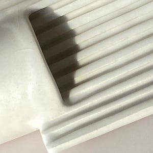 Pu&ograve; includere: Primo piano di un oggetto in ceramica bianca e scanalata. L'oggetto ha un angolo arrotondato e una superficie liscia e curva. La texture scanalata crea ombre, aggiungendo profondit&agrave; al design. L'illuminazione evidenzia la forma e la texture dell'oggetto.