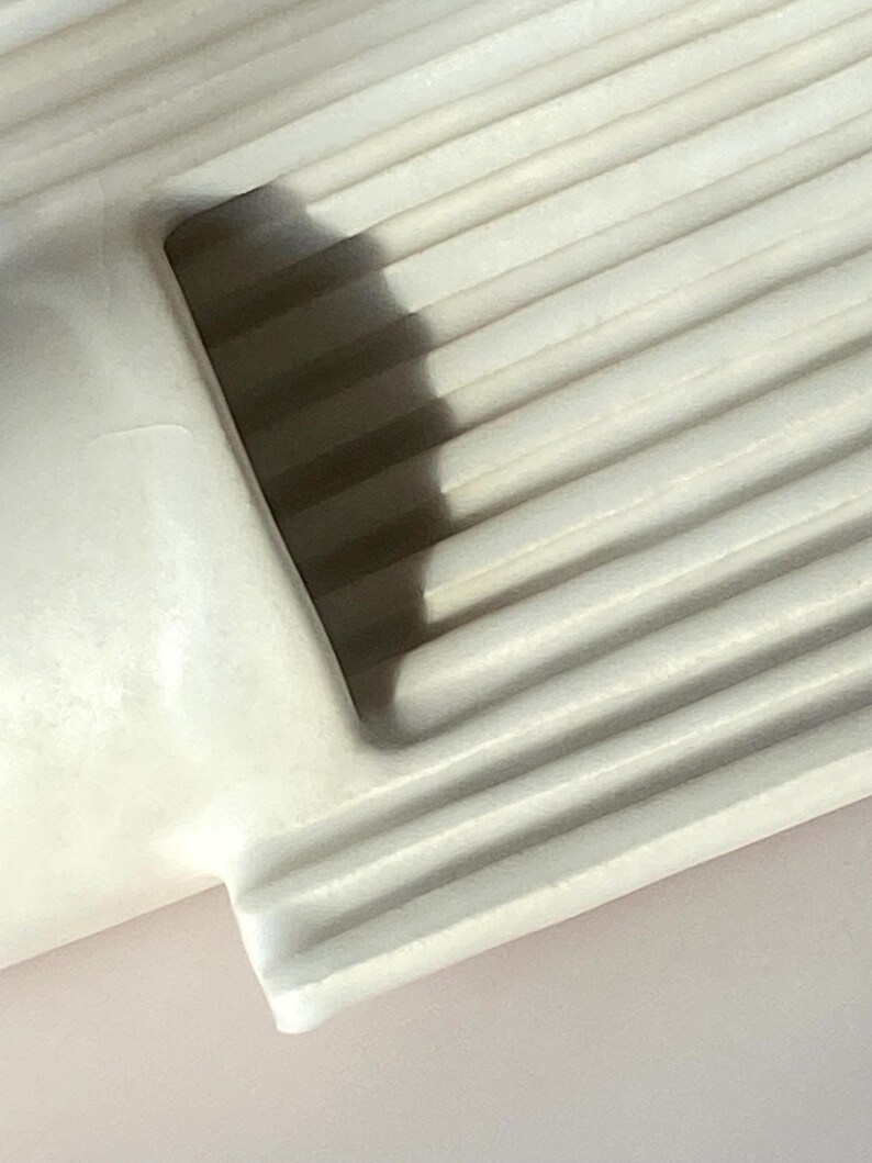 Pu&ograve; includere: Primo piano di un oggetto in ceramica bianca e scanalata. L'oggetto ha un angolo arrotondato e una superficie liscia e curva. La texture scanalata crea ombre, aggiungendo profondit&agrave; al design. L'illuminazione evidenzia la forma e la texture dell'oggetto.