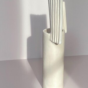 Pu&ograve; includere: Un vaso alto in ceramica bianco sporco con corpo cilindrico e un design unico e strutturato. La parte superiore presenta un elemento verticale a coste e un accento liscio e curvo. Il vaso &egrave; posto su uno sfondo bianco.