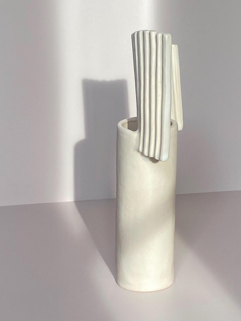 Pu&ograve; includere: Un vaso alto in ceramica bianco sporco con corpo cilindrico e un design unico e strutturato. La parte superiore presenta un elemento verticale a coste e un accento liscio e curvo. Il vaso &egrave; posto su uno sfondo bianco.