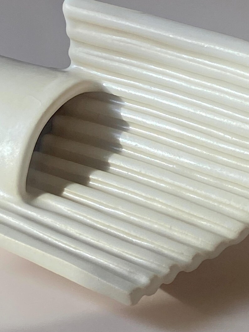 Pu&ograve; includere: Primo piano di un oggetto in ceramica color crema, a coste. L'oggetto ha un'apertura curva e cilindrica e una serie di creste parallele. Le creste creano una superficie strutturata e il design generale &egrave; semplice ed elegante.