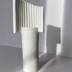 Pu&ograve; includere: Un vaso in ceramica bianca con base cilindrica e parte superiore rettangolare con scanalature verticali. Il vaso &egrave; posto su uno sfondo bianco, con ombre che aggiungono profondit&agrave;. Il design &egrave; moderno e minimalista.