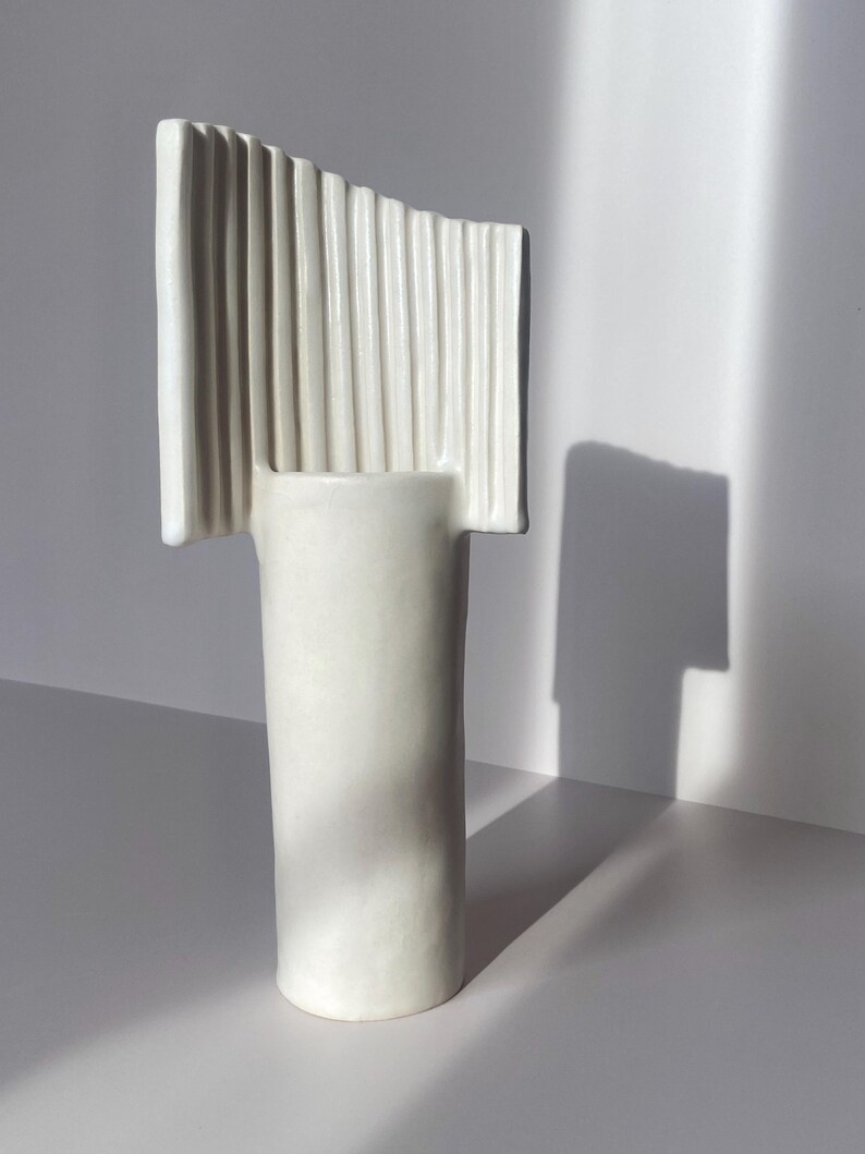Pu&ograve; includere: Un vaso in ceramica bianca con base cilindrica e parte superiore rettangolare con scanalature verticali. Il vaso &egrave; posto su uno sfondo bianco, con ombre che aggiungono profondit&agrave;. Il design &egrave; moderno e minimalista.