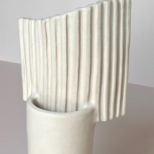 Pu&ograve; includere: Un vaso in ceramica color crema con base cilindrica e parte superiore curva a coste verticali. Il vaso presenta un design minimalista e una finitura liscia e opaca, adatta all'arredamento della casa.