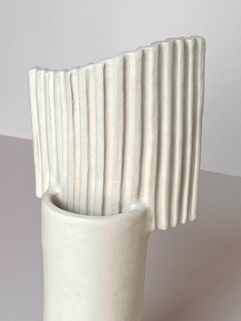 Pu&ograve; includere: Un vaso in ceramica color crema con base cilindrica e parte superiore curva a coste verticali. Il vaso presenta un design minimalista e una finitura liscia e opaca, adatta all'arredamento della casa.