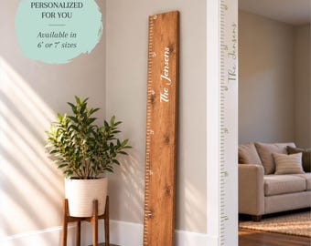 May include: A tall wooden growth chart ruler with a natural wood grain finish, featuring white measurement markings in feet and the personalized text The Jensens printed vertically in a white script font on the front surface.