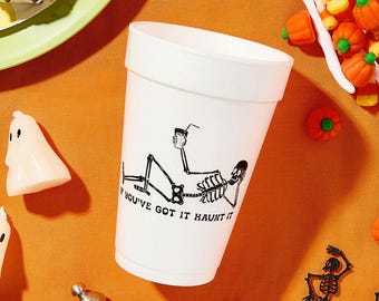 Puede incluir: Una taza de espuma blanca con un gráfico de esqueleto y el texto "IF YOU'VE GOT IT HAUNT IT" está rodeada de decoraciones temáticas de Halloween. Caramelos de maíz, caramelos de calabaza y decoraciones de esqueletos están esparcidos sobre una superficie naranja.