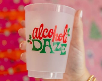 May include: A white plastic cup with the text "alcohol daze" in red and green lettering. The cup is being held by a hand wearing a gold bracelet.