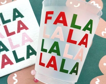 May include: A white cup with the words "FALA LALA LALA LALA" written in red, green, and pink. The cup is sitting on a green surface with gingerbread cookies.