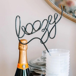May include: A clear glass bottle of Veuve Clicquot champagne with a gold label and a black wire 'boozy' sign on top. A stack of clear plastic cups with the text 'Toasting with the Talley's' printed on the side.