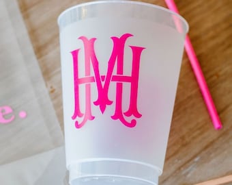May include: A white plastic cup with a pink monogram "MH" on the front. The cup is sitting on a wooden surface next to a pink plastic straw and a pink neon sign that says "you".