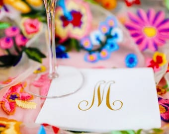 May include: A white cocktail napkin with a gold letter 'M' monogram on a colorful floral background. The napkin is next to a martini glass with a pink flower and ice.