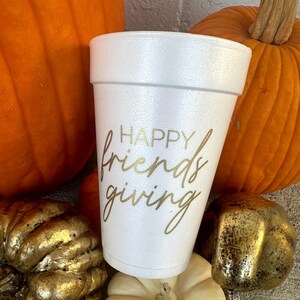 May include: A white foam cup with the words "HAPPY friends giving" in gold script. The cup is surrounded by orange pumpkins and gold-painted gourds, suggesting a fall or Thanksgiving theme.
